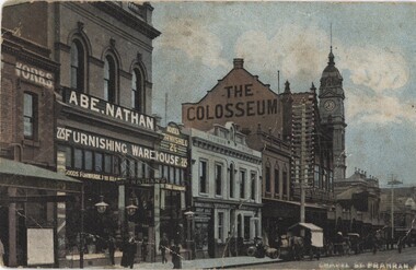 A row of early 1900s buildings along a street. There is a clocktower towards the end of the row and people walking along the footpath.