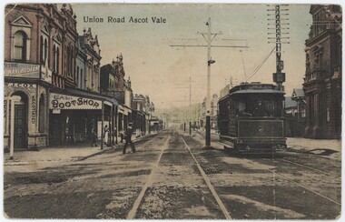 A street with two sets of tram tracks lined with 1890s-1900s buildings, with a person crossing the street and a green tram to the right.