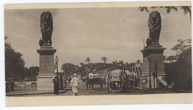 A postcard depicting two lion statues at the entrance to a bridge, as people and carts travel across.