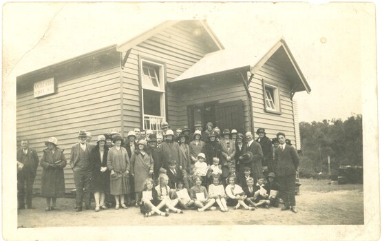 A group of standing adults and sitting children in front of the Macclesfield State School building.
