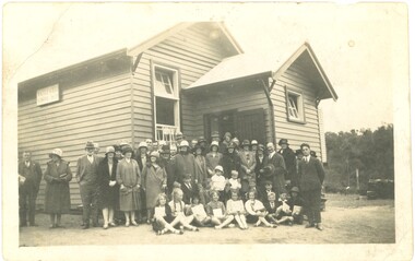 A group of standing adults and sitting children in front of the Macclesfield State School building.