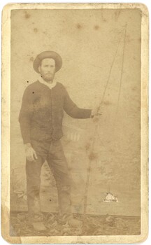 Photograph of man standing and facing the camera. He is wearing a hat and jacket and jeans. Sepia color.