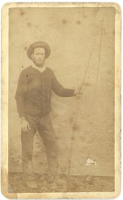Photograph of man standing and facing the camera. He is wearing a hat and jacket and jeans. Sepia color.