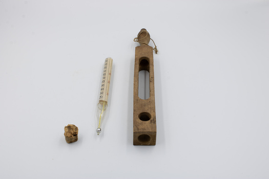 A thermometer, its empty wooden case and a cork laid out next to each other.