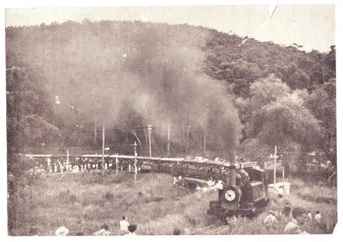 Black and white photograph of event in 1954 to mark Puffing Billy's last trip. The train crowded with people and with many well-wishers standing beside the track is crossing the road at Upper Ferntree Gully heading towards Belgrave.