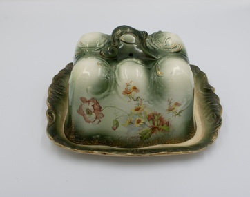 Ceramic cheese cover and plate with ornate design in green and white with gold highlights and floral decoration around all sides.