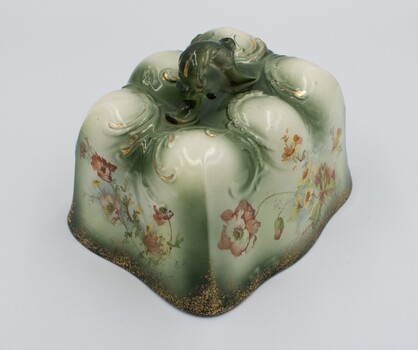 Two sides of rectangular ceramic cheese cover showing ornate design with floral decoration on the sides and handle on the top.