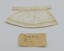 Front view of cream-colored doll's apron with original handwritten paper of maker's details