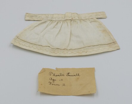 Front view of cream-colored doll's apron with original handwritten paper of maker's details