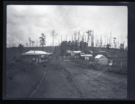 Black and white negative - landscape view - showing the main road in Gembrook heading west. The last building on the left is the Ranges Hotel and the second last building on the right is the double-gabled General Store which fronts the railway station.