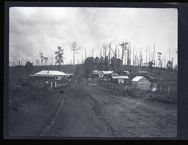 Black and white negative - landscape view - showing the main road in Gembrook heading west. The last building on the left is the Ranges Hotel and the second last building on the right is the double-gabled General Store which fronts the railway station.