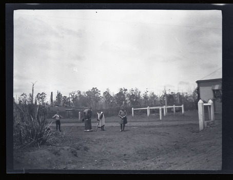 Black and white negative - landscape view - showing two boys and two girls playing backyard cricket. One boy is crouched on the 'stumps' which are an upturned pipe. One girl is the bowler, and the other girl is the batter.