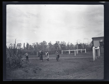 Black and white negative - landscape view - showing two boys and two girls playing backyard cricket. One boy is crouched on the 'stumps' which are an upturned pipe. One girl is the bowler, and the other girl is the batter.