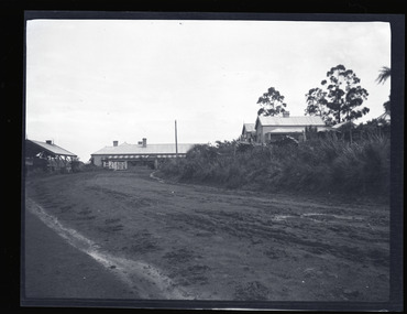 Black and white negative - landscape view - showing a road running alongside the railway station towards the Ranges Hotel, Gembrook with the General Store visible on the right and railway gates in front of the hotel.
