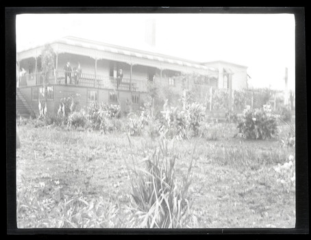 Black and white landscape view photograph of a side and back view of the Ranges Hotel, Gembrook with three men in suits and a woman standing on the balcony.