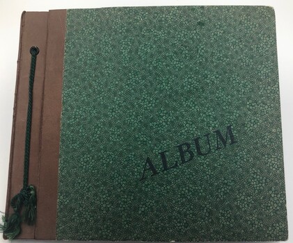 Front cover of postcard album. Embossed green. Brown leather binding with green cord.