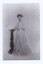 Formal, studio black and white photograph depicting a young woman in a floor length white evening gown posing next to a black pedestal.
