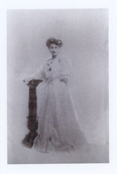 Formal, studio black and white photograph depicting a young woman in a floor length white evening gown posing next to a black pedestal.
