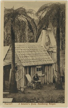 Wooden house surrounded by trees with man sitting in front, captured in black and white but somewhat yellowed.
