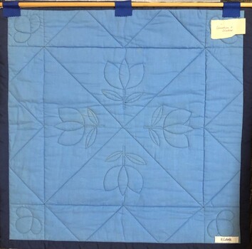 Back of patchwork quilt showing wooden dowel fixed at the top for hanging and handwritten notes attached to the right top and the right bottom of the quilt.
