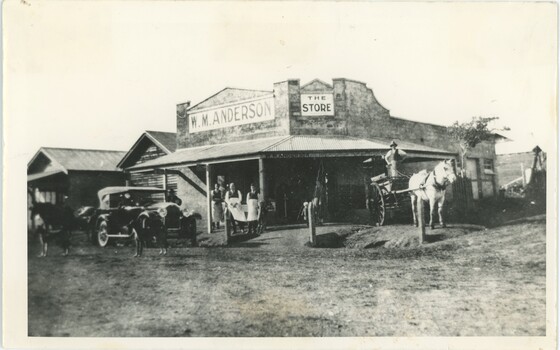 A black and white photograph depicting a corner store with a car, horses, and people in front.