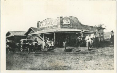 A black and white photograph depicting a corner store with a car, horses, and people in front.