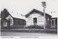 A black and white photograph featuring two buildings by a road, with publication and photograph details printed below.