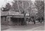 The obverse of a black and white photograph, featuring a front-left view of a wooden building with trees and a road behind.