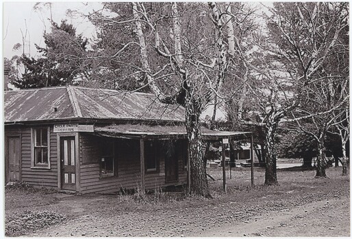 The obverse of a black and white photograph, featuring a front-left view of a wooden building with trees and a road behind.