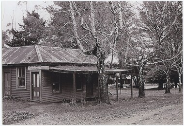 The obverse of a black and white photograph, featuring a front-left view of a wooden building with trees and a road behind.