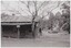 The obverse of a black and white photograph, featuring a front view of a wooden building with flat ground and trees behind.