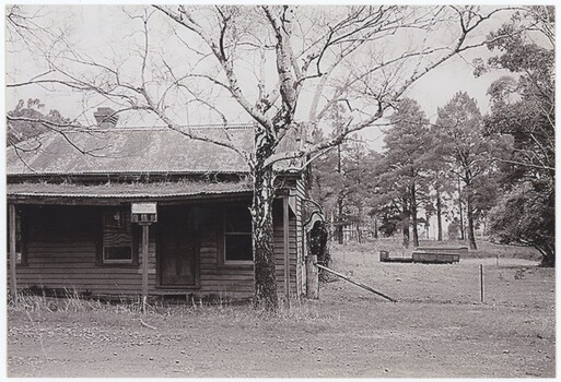 The obverse of a black and white photograph, featuring a front view of a wooden building with flat ground and trees behind.