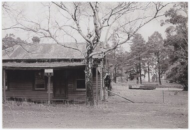 The obverse of a black and white photograph, featuring a front view of a wooden building with flat ground and trees behind.