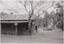 The obverse of a black and white photograph, featuring a front view of a wooden building with flat ground and trees behind.