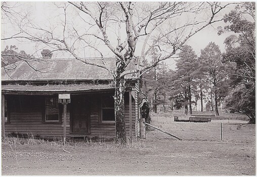 The obverse of a black and white photograph, featuring a front view of a wooden building with flat ground and trees behind.