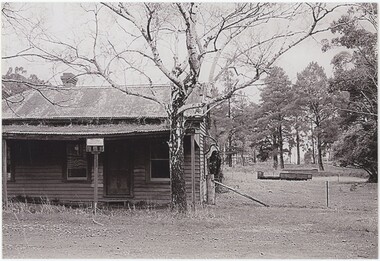 The obverse of a black and white photograph, featuring a front view of a wooden building with flat ground and trees behind.