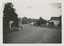 A black and white photograph of a dirt road with a parked car and buildings.