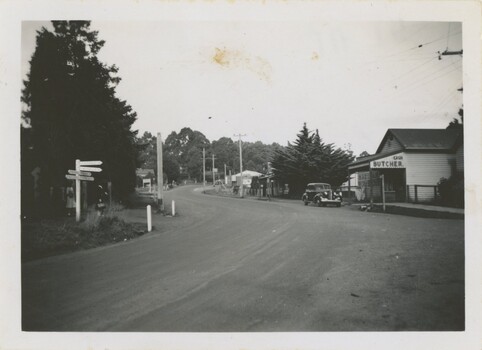 A black and white photograph of a dirt road with a parked car and buildings.