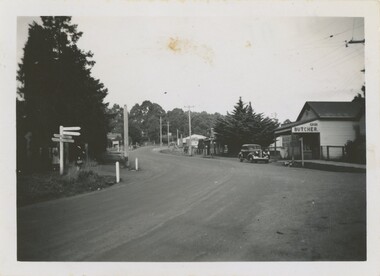 A black and white photograph of a dirt road with a parked car and buildings.
