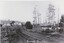 A black and white photograph depicting an unsealed road lined by occasional wooden structures. A label is in the centre-bottom of the image.