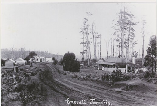 A black and white photograph depicting an unsealed road lined by occasional wooden structures. A label is in the centre-bottom of the image.