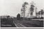 A black and white photograph depicting an unsealed road lined by occasional wooden structures. A label is in the centre-bottom of the image.