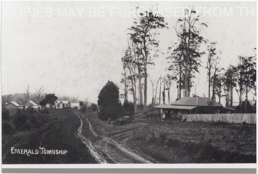 A black and white photograph depicting an unsealed road lined by occasional wooden structures. A label is in the centre-bottom of the image.