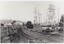 A black and white photograph depicting an unsealed road lined by occasional wooden structures. A label is in the centre-bottom of the image.