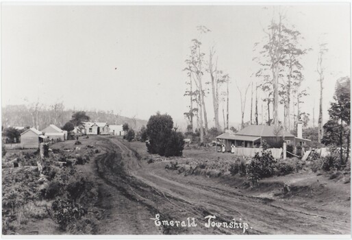 A black and white photograph depicting an unsealed road lined by occasional wooden structures. A label is in the centre-bottom of the image.