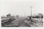 A printed black and white photograph depicting an unpaved road with a store on the right and several other buildings on the left.