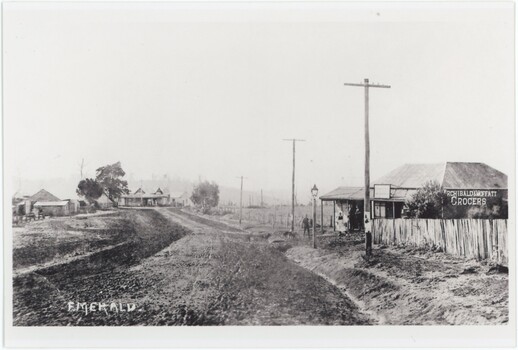 A printed black and white photograph depicting an unpaved road with a store on the right and several other buildings on the left.