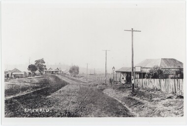 A printed black and white photograph depicting an unpaved road with a store on the right and several other buildings on the left.