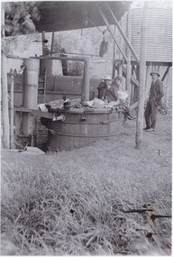 A black and white photograph of a eucalyptus distillery, with two women sitting on a storage tank and a man standing beside them.