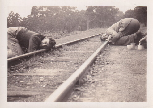Two young men lying down next to the railway line listening for sound on the rails.